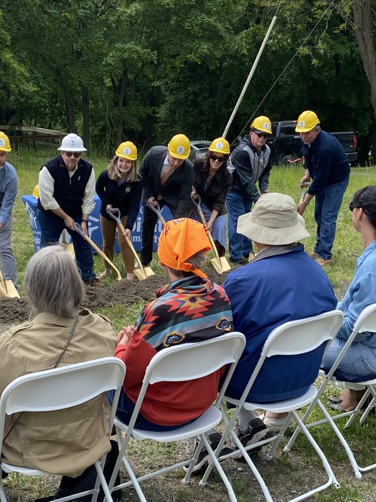 Seven people in hard hats pointing golden shovels into a mound of dirt with people sitting in seats before them.